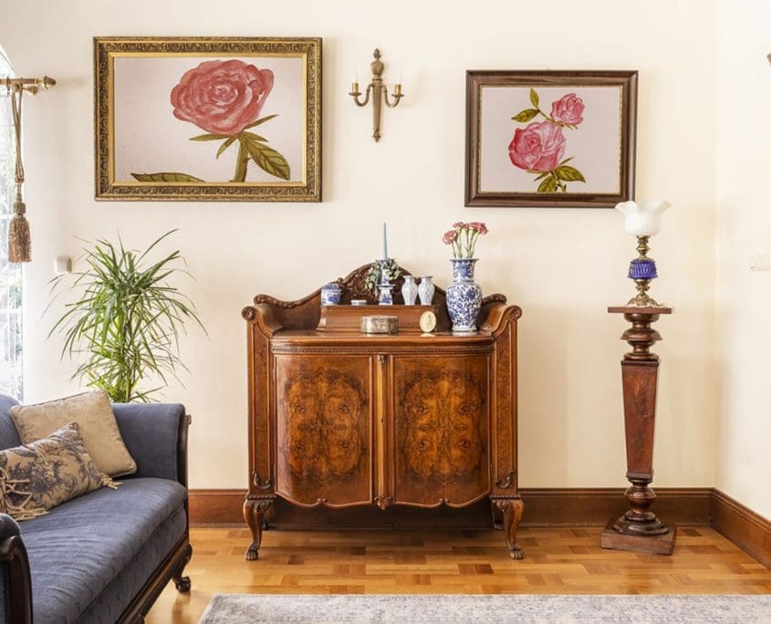 In this stylish living room curated by the Asset Valuations Group, a classic wooden sideboard is elegantly decorated with blue and white vases, complemented by a lamp on a stand. Two rose paintings grace the wall above, while a plush blue couch with cushions rests on the polished wooden floor to the left.