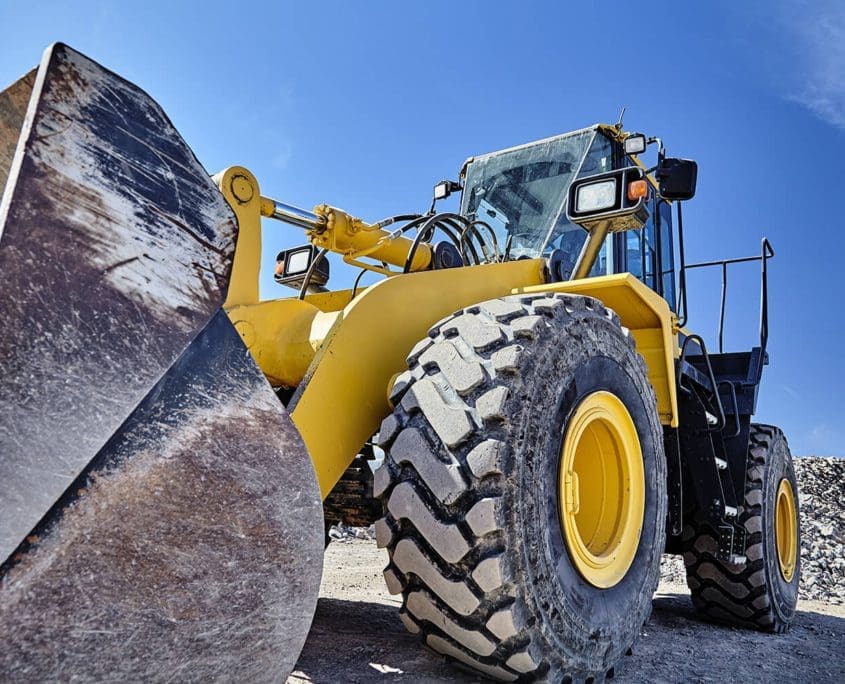 A yellow front-end loader with large tires and a scoop stands diagonally against a blue sky at an Asset Valuations Group construction site, surrounded by gravel piles.