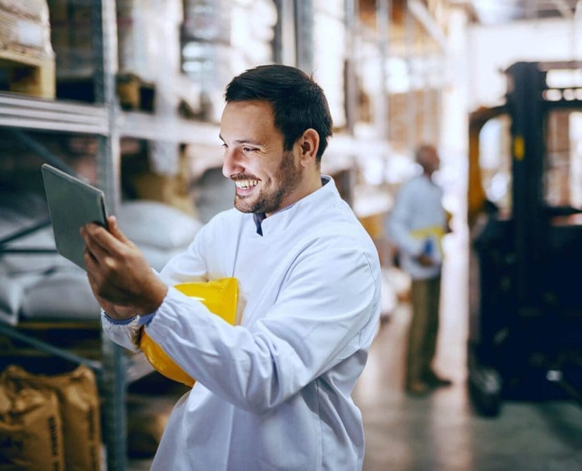 A man in a white coat, representing the Asset Valuations Group, smiles at a tablet while holding a yellow hard hat. He stands in a warehouse with shelves filled with large bags. Another person is blurred in the background near shelving.