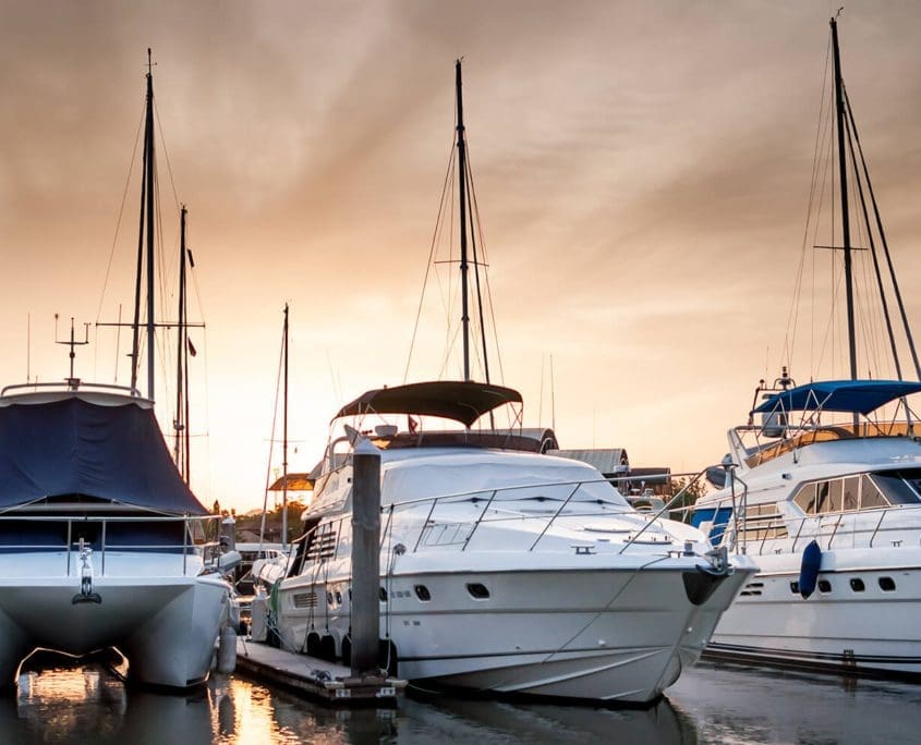 As the sun dips below the horizon, casting warm orange and yellow hues over the marina, the Asset Valuations Group surveys the scene where docked yachts and sailboats rest. Tall masts reach into the sky, capturing this serene setting reflected softly on tranquil waters.