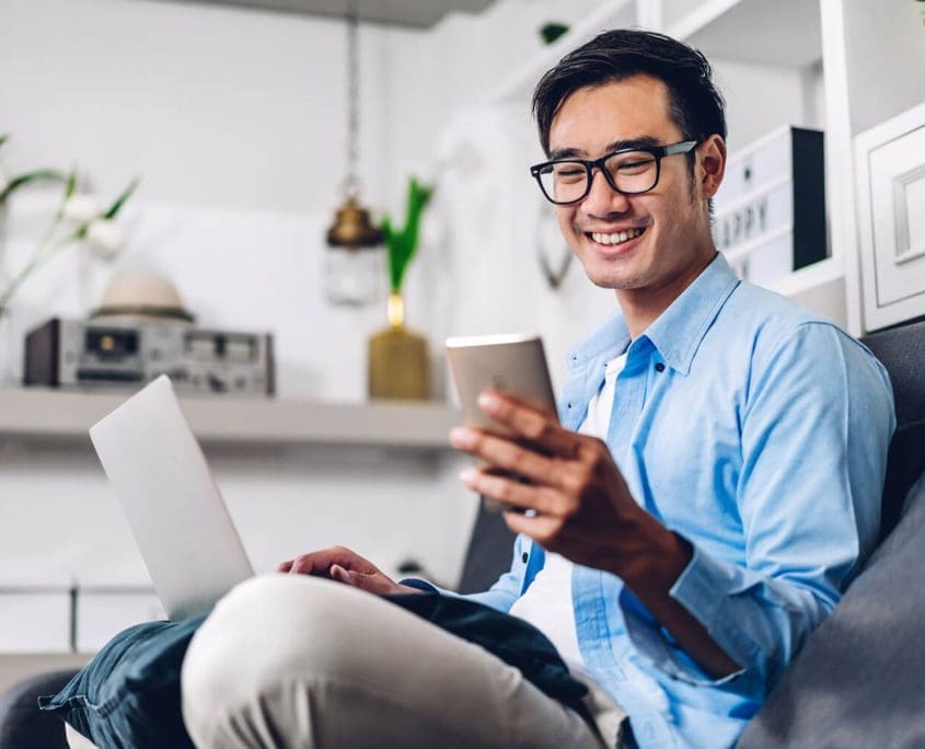 A man in a blue shirt and glasses is sitting on a couch, smiling at his phone. He has a laptop on his lap, potentially reviewing notes for the Asset Valuations Group. The background features shelves with decor and a retro radio.