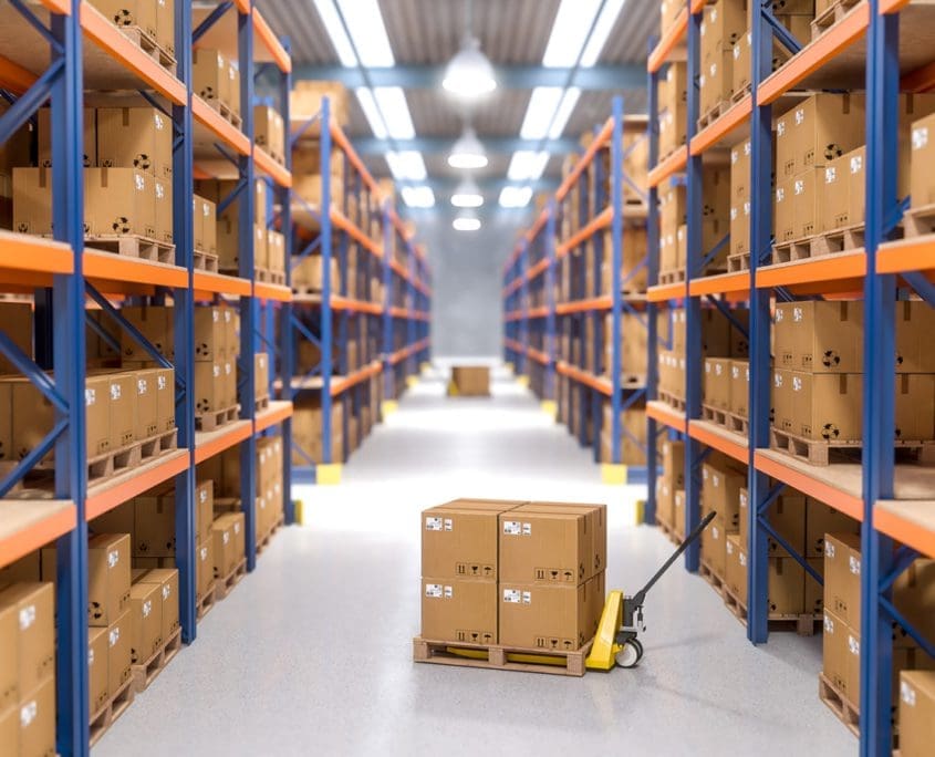Wide view of an organized warehouse aisle lined with tall blue and orange shelves filled with cardboard boxes. In the foreground, a hand pallet truck holds two boxes under bright overhead lighting, ensuring everything is in perfect order for the Asset Valuations Group.