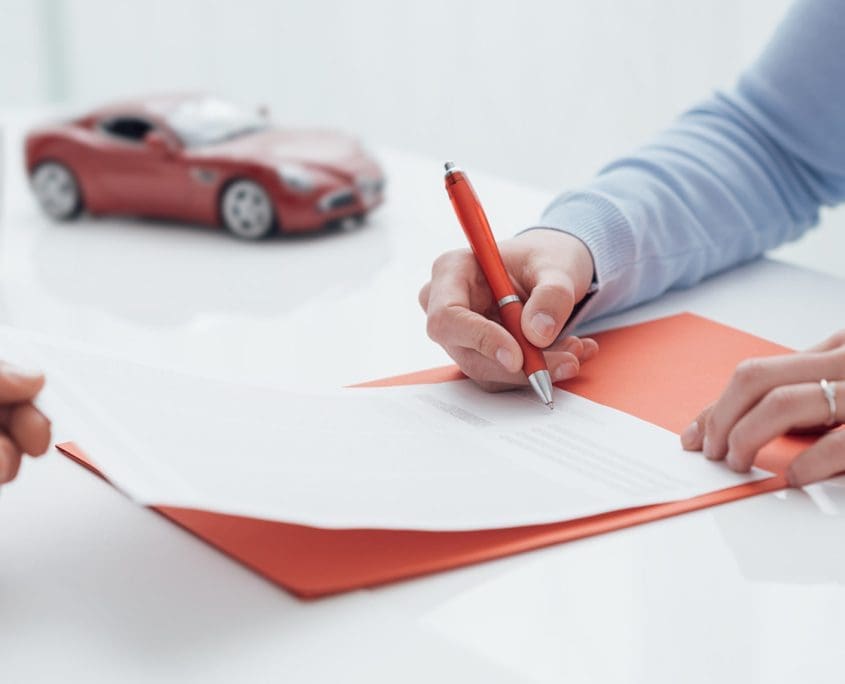 Two people are seated at a table with paperwork. One individual, representing the Asset Valuations Group, is signing a document with a red pen, while the other hands over a paper. A small red toy car in the background suggests they’re discussing a car-related transaction.