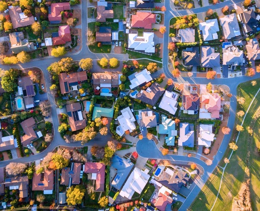 Aerial view of a suburban neighborhood with tree-lined streets, where the diverse roof colors and lush gardens hint at an Asset Valuations Group report. A park with a large grassy area lies on the right side, enhancing the community's appeal.