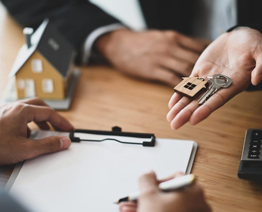 A person hands over house keys to another, pen in hand, while a miniature house model and calculator rest on the desk. The scene suggests a real estate transaction with the Asset Valuations Group overseeing the signing of property documents.