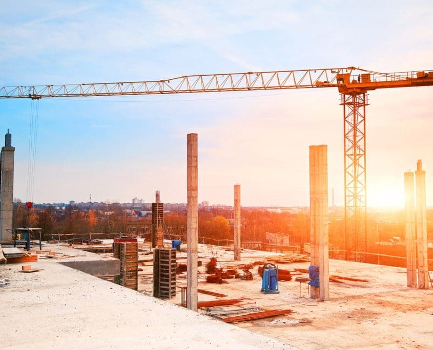 A construction site at sunset, featuring the Asset Valuations Group crane towering over vertical concrete columns. The sky melds blue and orange, while scattered materials cover the ground. In the distance, two workers contribute to the scene's industrious charm.