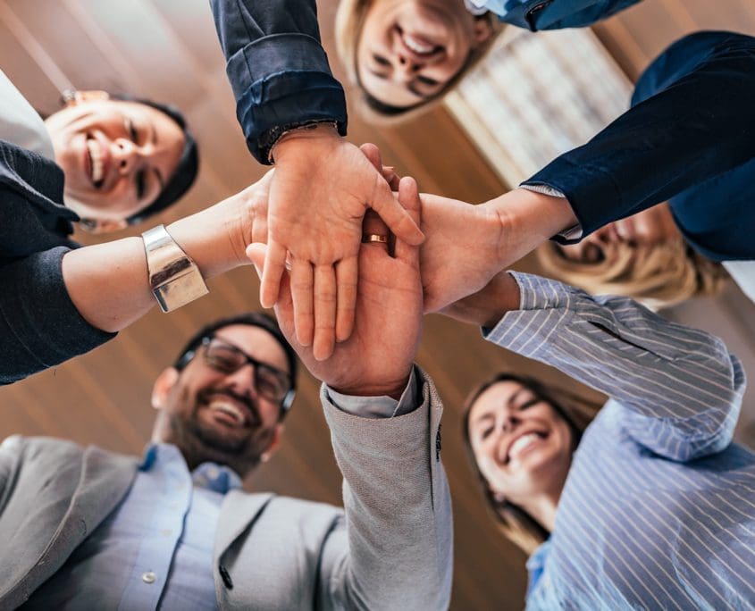 Team building, partnership, business success concept. Bottom vie A group of five people, part of the Asset Valuations Group, stand in a circle viewed from below. They are smiling and stacking their hands together in the center, showcasing teamwork and unity against a backdrop of wooden-paneled ceilings.