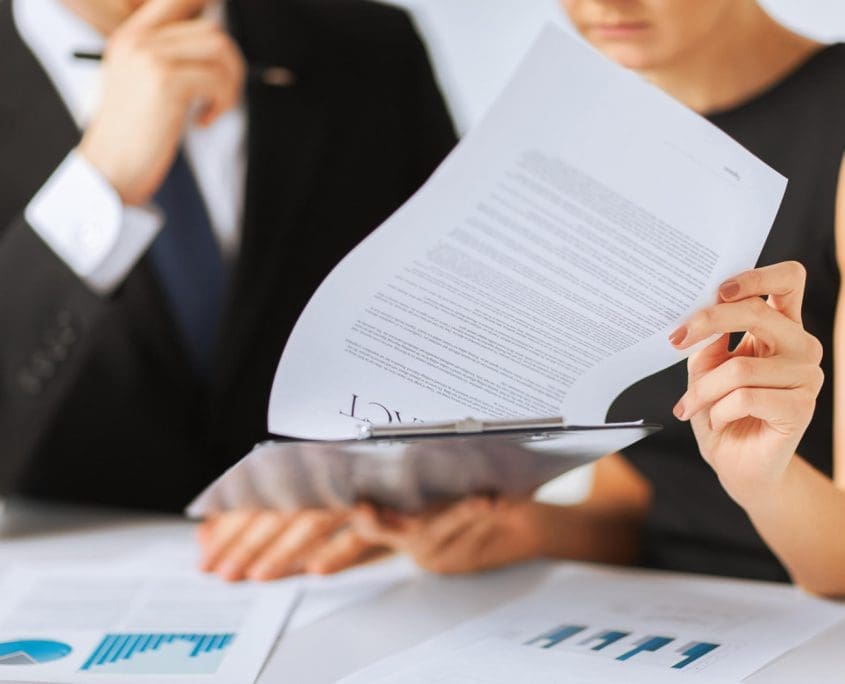 Two people in business attire, from the Asset Valuations Group, are reviewing documents. One holds a report with charts and text, while the other has their hand on their chin in thought. More charts are on the table in front of them.