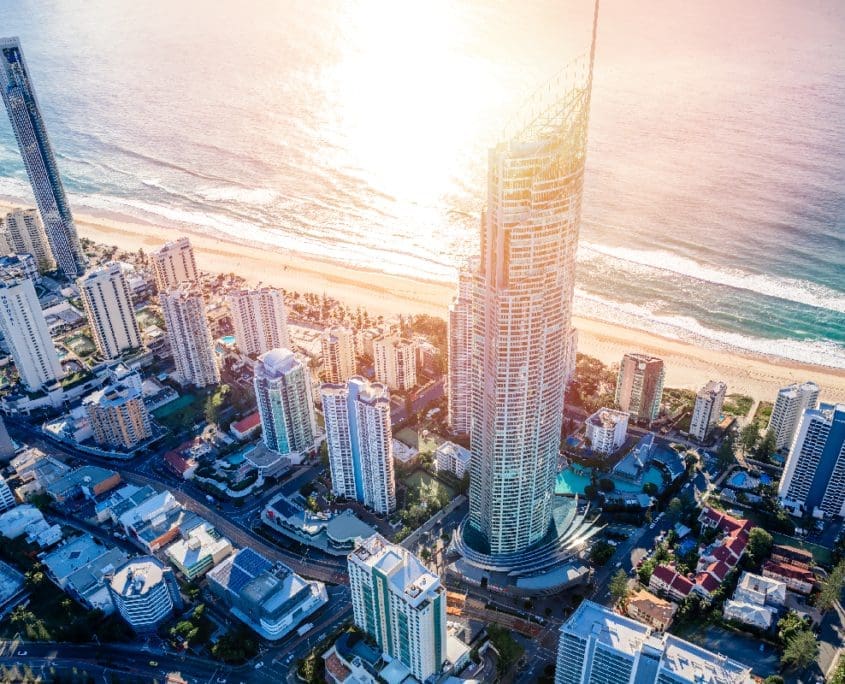Aerial view of a vibrant coastal cityscape with high-rise buildings near a sandy beach. The sun glows brightly over the ocean, casting a warm light across the scene, as the Asset Valuations Group assesses this bustling urban environment and serene seashore.