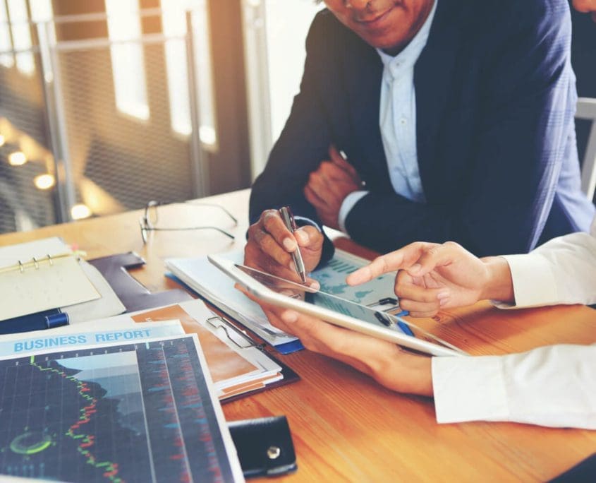 Two people from the Asset Valuations Group are reviewing a tablet at a desk cluttered with papers, charts, and a business report. One person holds a pen, pointing at the tablet, while the other gestures to the screen. Both are in business attire in a well-lit office.