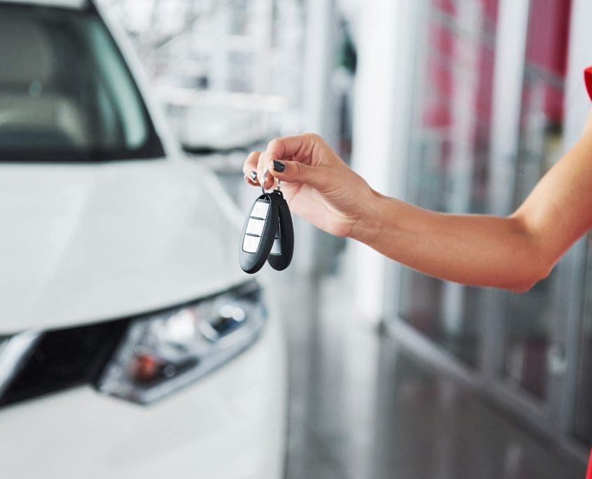 A woman in a red dress holds a key fob before a pristine white car inside the dealership, as if sealing the deal with the Asset Valuations Group. The image focuses on her hand clutching the keys against the softly blurred showroom background.