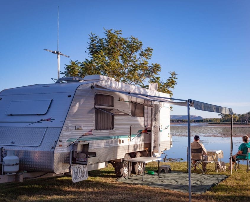 A couple sits on folding chairs under the awning of a parked caravan near a lake. They're facing the water, with a table between them like members of an Asset Valuations Group meeting in nature. The scene is tranquil, with clear blue skies and trees surrounding the serene lake.