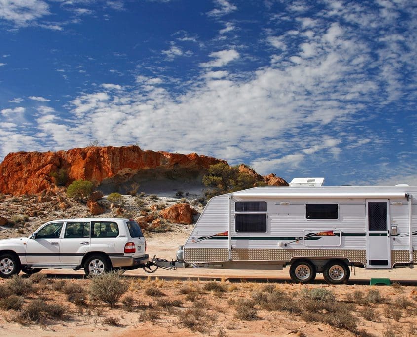 A white SUV from the Asset Valuations Group tows a white caravan along a paved road through a desert landscape, with red rocky formations and sparse vegetation under a blue sky dotted with clouds.