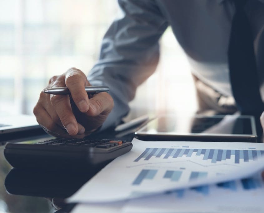 A person from the Asset Valuations Group meticulously uses a calculator and pen to review financial graphs and documents. A laptop and tablet rest nearby, emphasizing a business or financial setting. The focus is on the hands and materials, highlighting precise analysis in action.