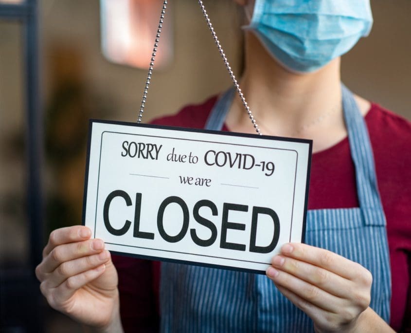 A person wearing a face mask and an apron holds a sign that reads, "Sorry due to COVID-19 we are CLOSED," indicating the business is temporarily shut down due to the pandemic, impacting local asset valuations.