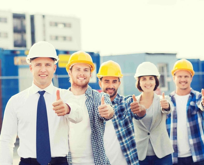 Five construction workers, representing the Asset Valuations Group, stand outdoors donning hard hats and a mix of casual or business attire. With buildings and shipping containers in the backdrop, they are smiling and giving a thumbs-up gesture, exemplifying teamwork and expertise.