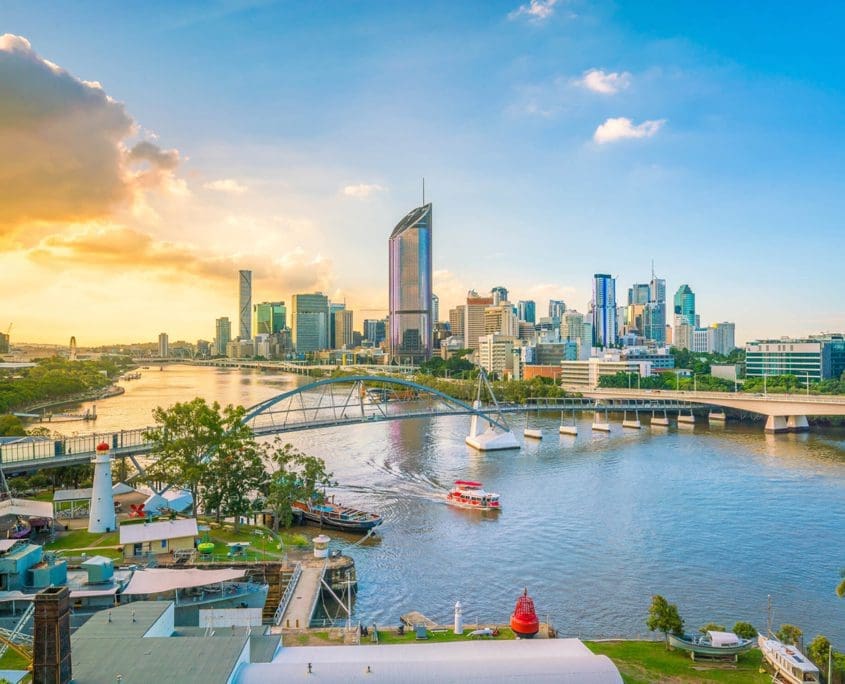A vibrant cityscape features a river with boats, a bridge, and a skyline of modern skyscrapers under a partly cloudy sky during sunset. In the foreground, the Asset Valuations Group's offices add to the dynamic waterfront area filled with trees and buildings.