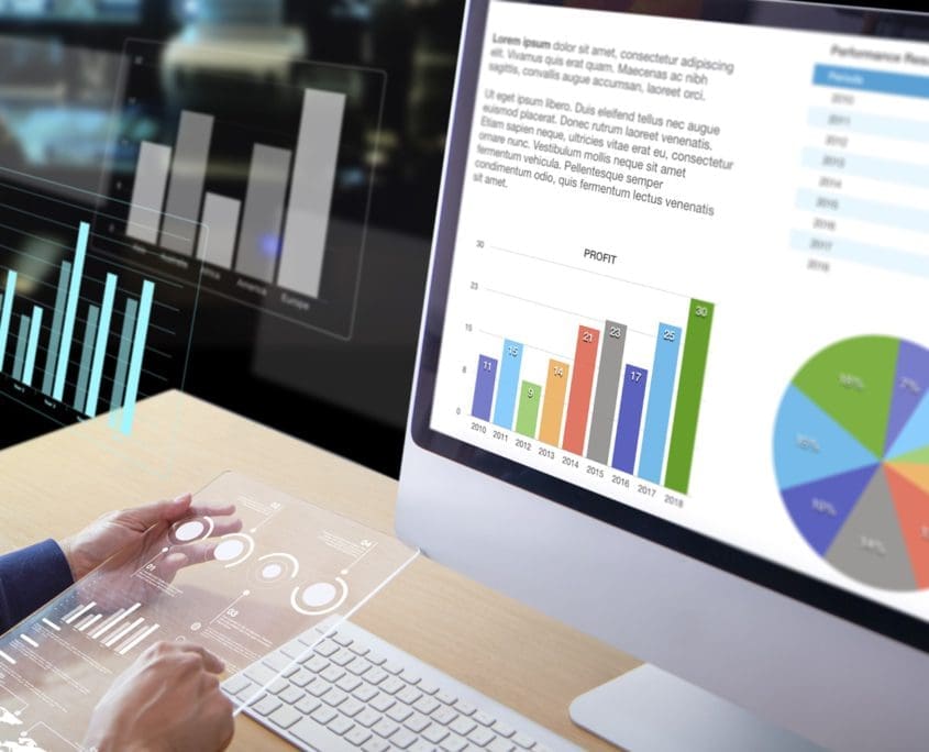 A person examines data on a computer screen displaying various charts, including bar graphs, a pie chart, and text summaries. Nearby, a transparent tablet shows additional Asset Valuations Group insights. The scene suggests data analysis or business reporting.