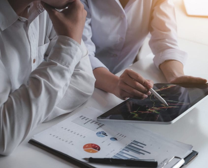 Two people in white shirts from the Asset Valuations Group analyze a graph on a tablet. One holds a stylus pointing at the screen, while a printed chart with bar graphs and pie charts lies on the desk. The brightly lit scene suggests a professional setting.