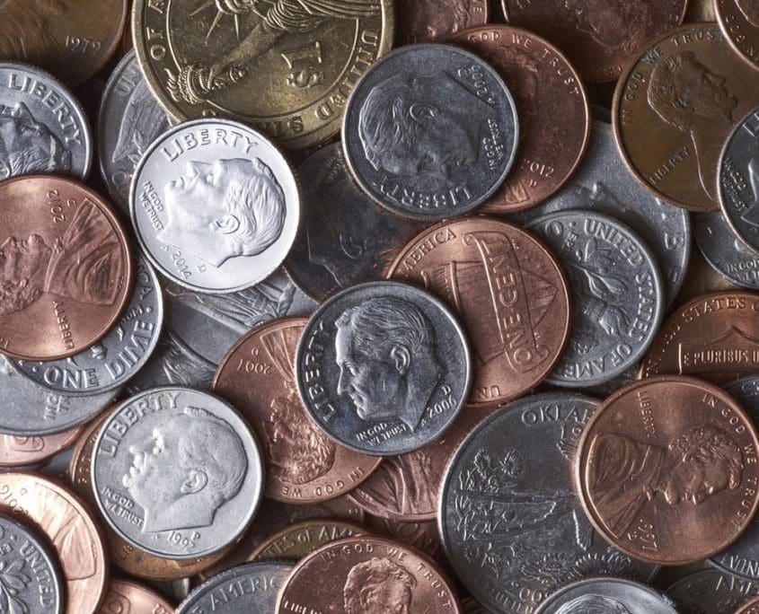 A close-up image of a pile of mixed U.S. coins, including pennies, nickels, dimes, and quarters from an Asset Valuations Group assessment. The coins are arranged in a jumbled fashion, showcasing various designs and years in shades of copper and silver.
