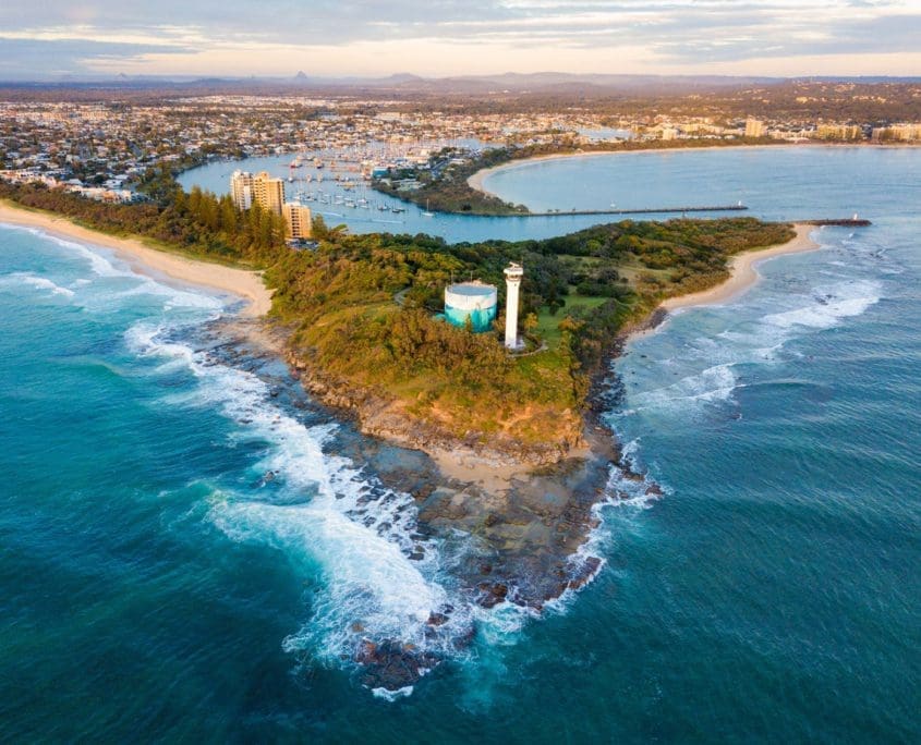 Aerial view of a coastal landscape featuring a rocky headland with a lighthouse overlooks a strategic location assessed by Asset Valuations Group. The surrounding area includes beaches, greenery, and a curved coastline leading to a town vibrant with buildings and boats in the marina.