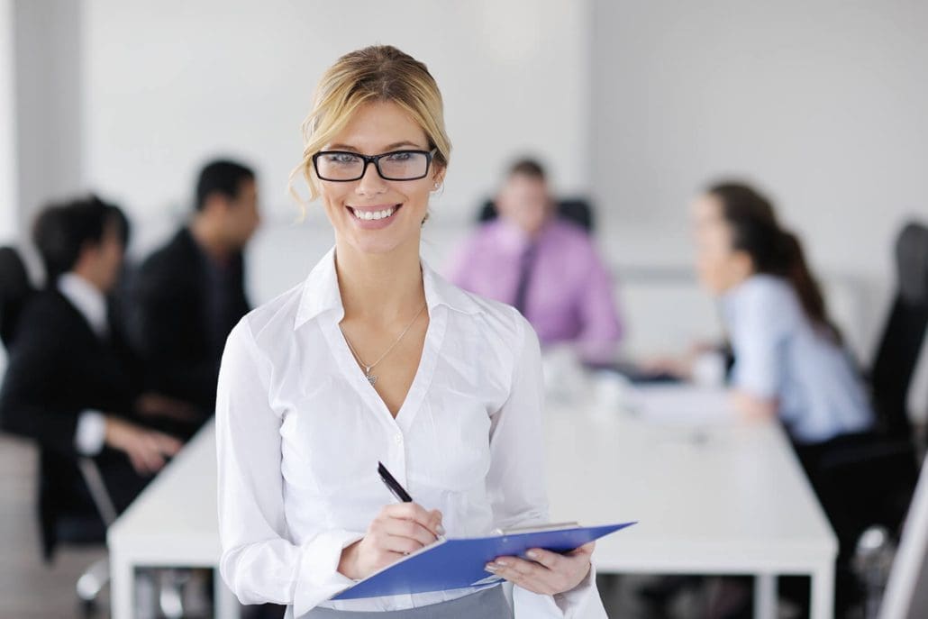 A smiling woman with glasses and a white blouse holds a blue clipboard and pen, representing the Asset Valuations Group in an office. She stands in focus while four blurred colleagues sit talking around a conference table in the background.