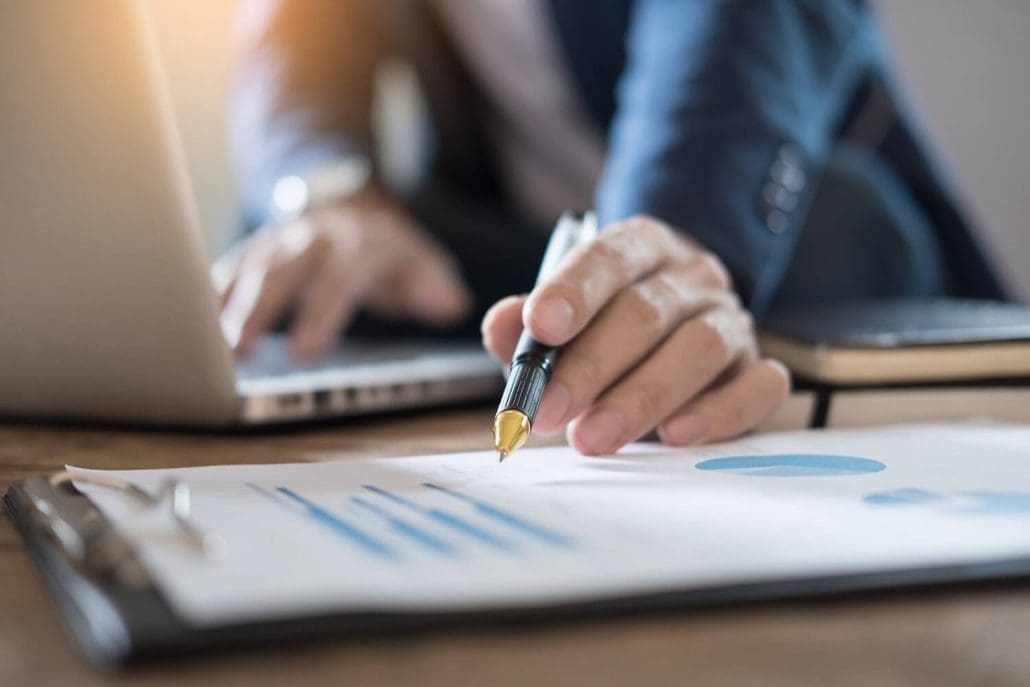 A person in a suit works at a desk with a laptop, meticulously analyzing data for the Asset Valuations Group. They hold a pen, pointing at charts and graphs on a clipboard, ensuring each valuation is precise and accurate.