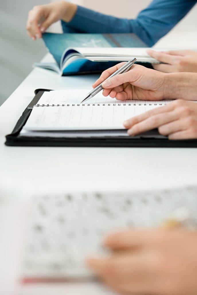 Close-up of a person from the Asset Valuations Group writing in a spiral-bound notebook with a pen on a white desk. Nearby, blurred pages of an open book hint at the focused study or work environment.