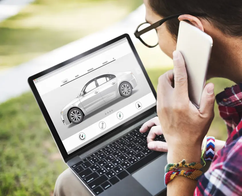 A person wearing glasses is using a laptop to view a car design application. They're on the phone with the Asset Valuations Group while examining the laptop screen, which displays a side view of a car along with various customization options below.