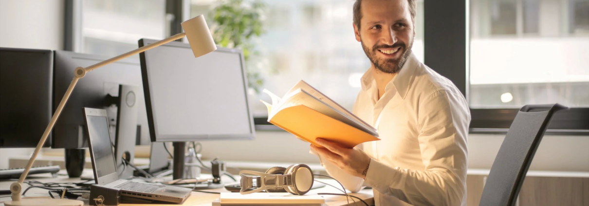 In the Asset Valuations Group office, a man in a white shirt sits comfortably in an office chair at his desk, surrounded by his computer and lamp. He's holding an open book and smiling as he glances to the side, with large windows offering a blurred cityscape view behind him.