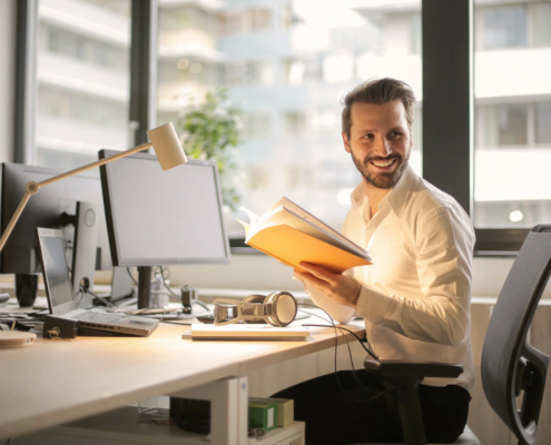 In the Asset Valuations Group office, a man in a white shirt sits comfortably in an office chair at his desk, surrounded by his computer and lamp. He's holding an open book and smiling as he glances to the side, with large windows offering a blurred cityscape view behind him.