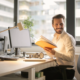 In the Asset Valuations Group office, a man in a white shirt sits comfortably in an office chair at his desk, surrounded by his computer and lamp. He's holding an open book and smiling as he glances to the side, with large windows offering a blurred cityscape view behind him.