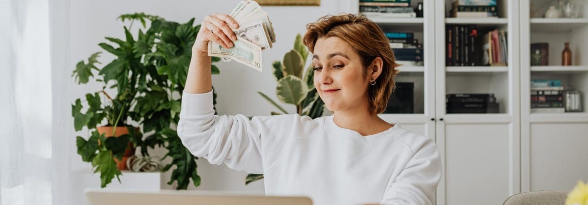 A person from the Asset Valuations Group sits at a desk, holding a fan of dollar bills with a satisfied expression. A laptop and glass rest on the table, while bookshelves filled with books and a potted plant adorn the background.