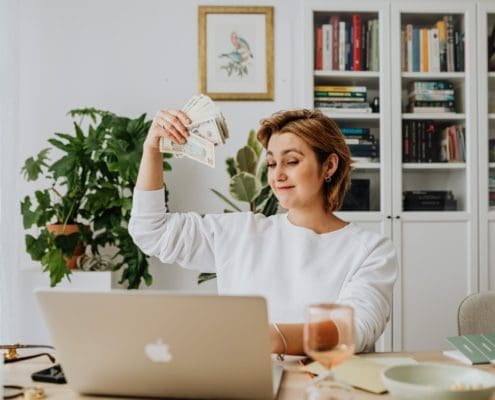 A person from the Asset Valuations Group sits at a desk, holding a fan of dollar bills with a satisfied expression. A laptop and glass rest on the table, while bookshelves filled with books and a potted plant adorn the background.