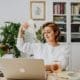 A person from the Asset Valuations Group sits at a desk, holding a fan of dollar bills with a satisfied expression. A laptop and glass rest on the table, while bookshelves filled with books and a potted plant adorn the background.