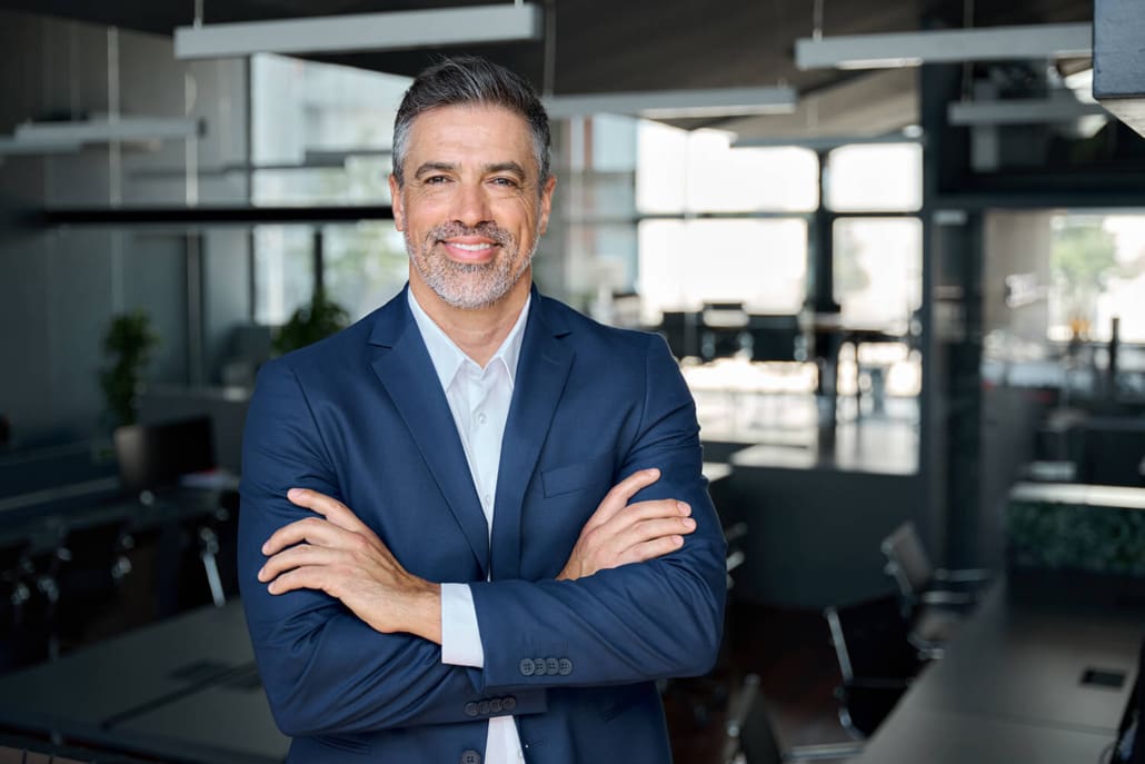 A confident man with salt-and-pepper hair and a beard, part of the Asset Valuations Group, smiles while standing with arms crossed, wearing a navy suit and white shirt. He is in a modern office space with large windows and glass partitions in the background.