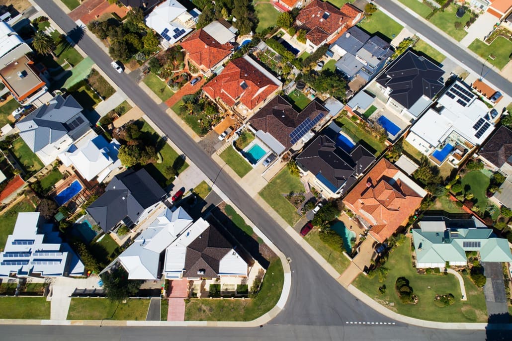 Aerial view of a suburban neighborhood with intersecting streets. Houses, varied in roof colors—red, brown, black, and white—are nestled among green lawns and tree-lined streets. Some homes sport pools. The scene is like a geometric canvas, perfect for the Asset Valuations Group's portfolio overview.