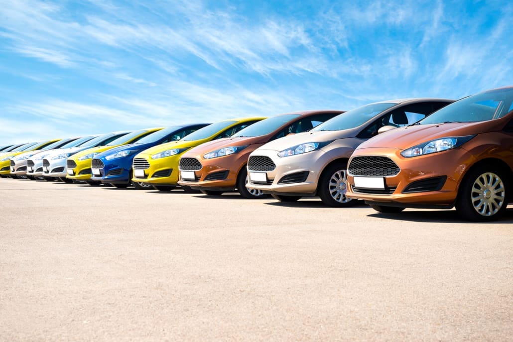 A row of colorful cars, arrayed like the finest assets in an Asset Valuations Group portfolio, is parked side by side on a concrete surface under a bright blue sky. The cars, featuring shades from white to orange, are aligned diagonally from the foreground to the background.