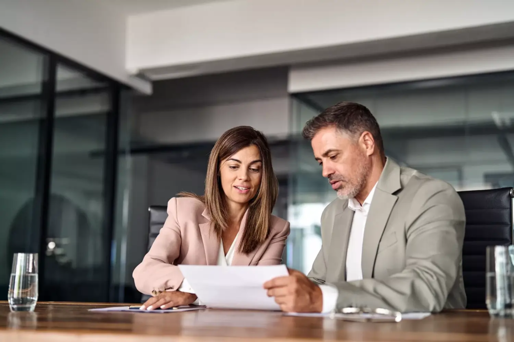 Two business professionals, a woman and a man, sit at a conference table in a modern office reviewing a document on What Are Asset Registers. Both are focused and engaged, with glasses of water on the table.