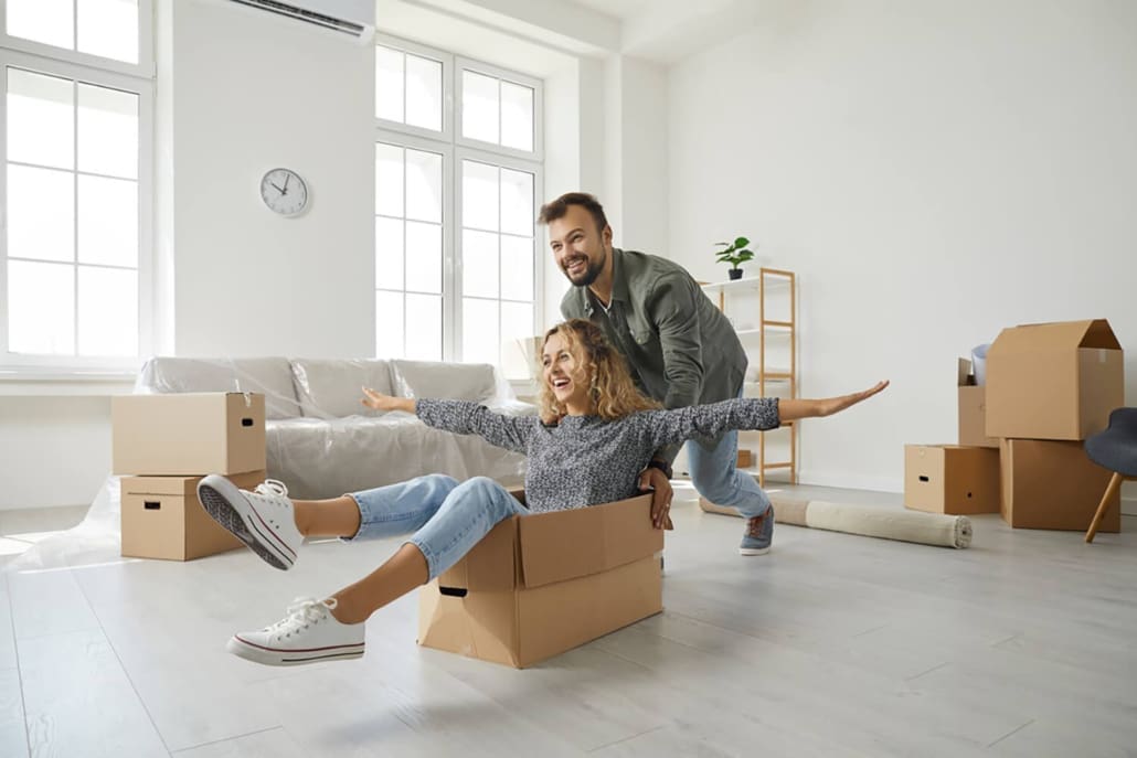 A smiling man pushes a laughing woman who is sitting in a cardboard box with her arms outstretched. Surrounded by moving boxes in a bright, unfurnished Sydney room, they share a joyful moment during their move.