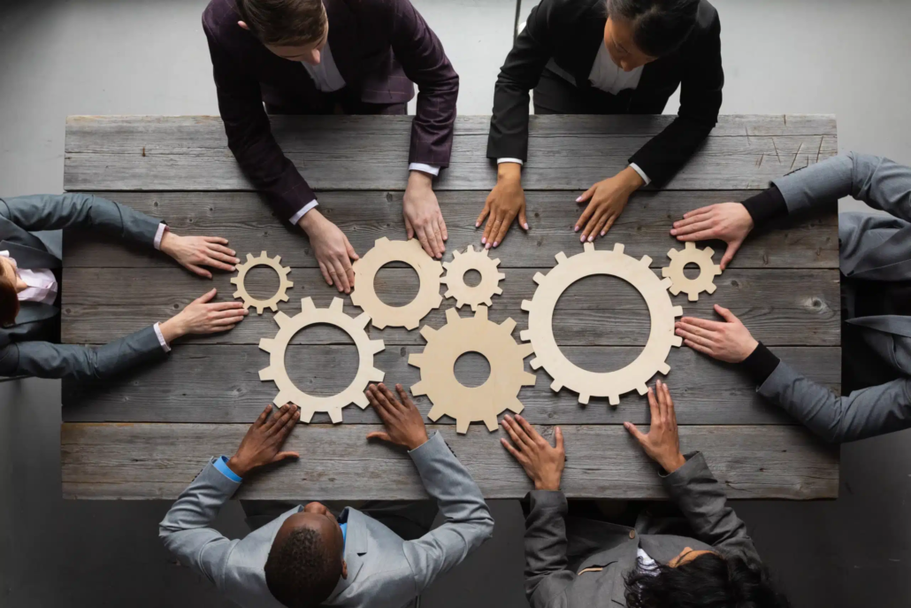 Six people in business attire sit around a wooden table, each reaching toward wooden gear shapes, symbolizing teamwork and collaboration essential for effective Purchase Price Allocation Valuation in a business or organizational setting.