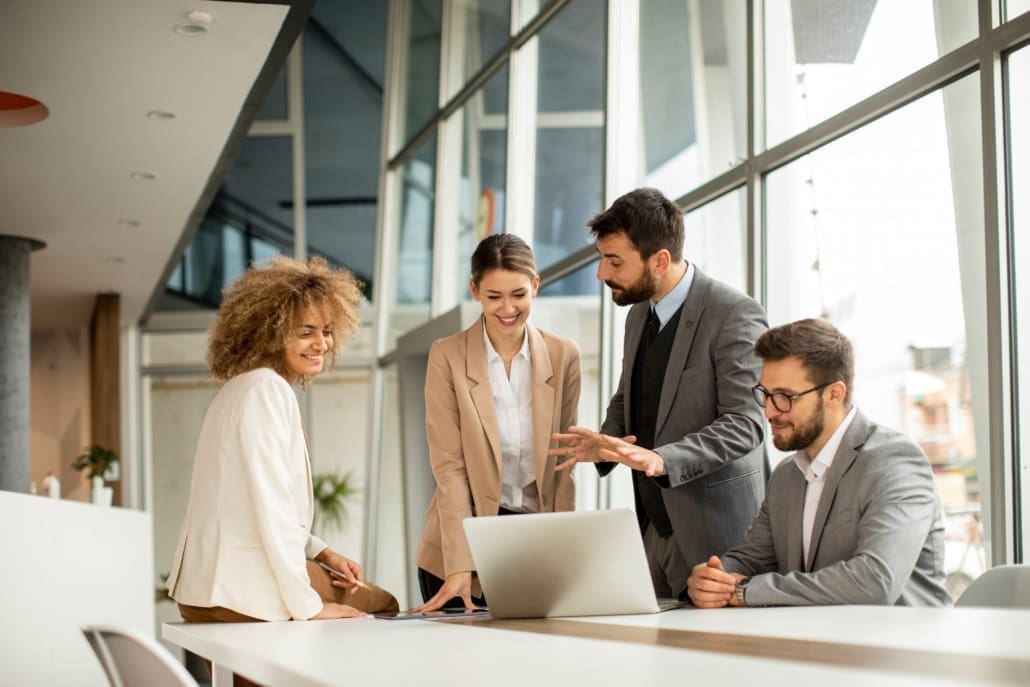 Purchase Price Allocation Valuation, Four business professionals, two women and two men, stand and sit around a laptop at a modern office table, smiling as they discuss Purchase Price Allocation Valuation in front of large windows.