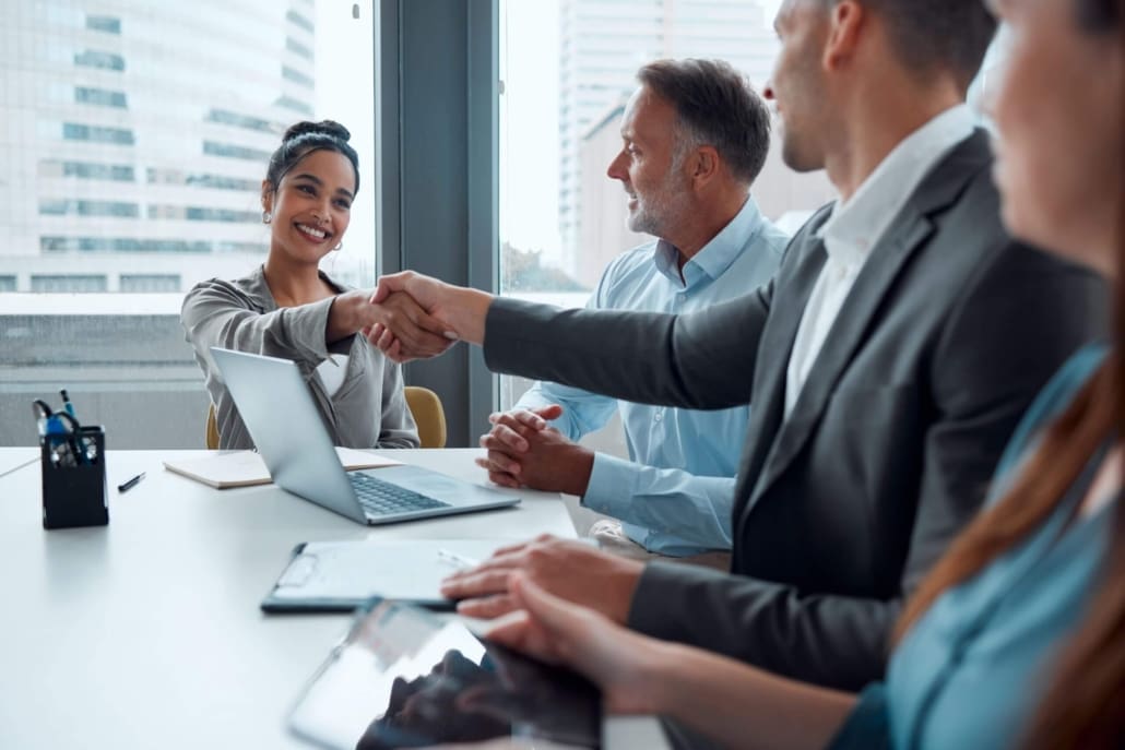 Four business professionals sit at a table in a modern office. Two, a woman and a man, are smiling and shaking hands, while the others look on—capturing the moment of agreement after discussing small business valuations.