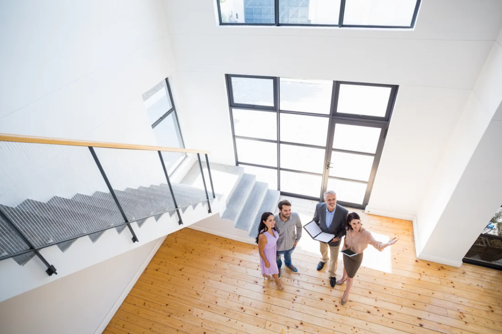 A group of four people stands in a modern, bright entryway with large windows and wooden floors, looking up toward the camera—a welcoming scene often seen in homes appraised by VALUATIONS BRISBANE. A staircase with a metal railing is visible on the left.