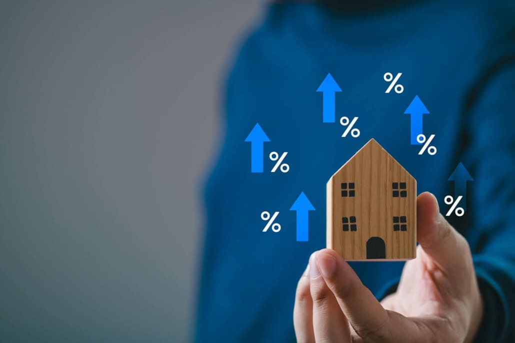 Property Valuer Online, A person holds a small wooden house model. Blue arrows and percent signs float upward around the house, symbolizing rising housing prices or increasing mortgage interest rates—a scenario where a property valuer online can help assess real-time values.