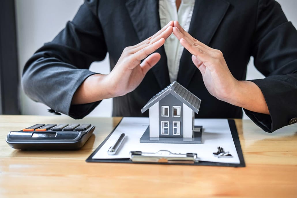 Property Valuers Sydney, A person in a suit covers a small model house with their hands, symbolizing protection. A calculator, clipboard, and keys are on the table, suggesting real estate services by Property Valuers Sydney.