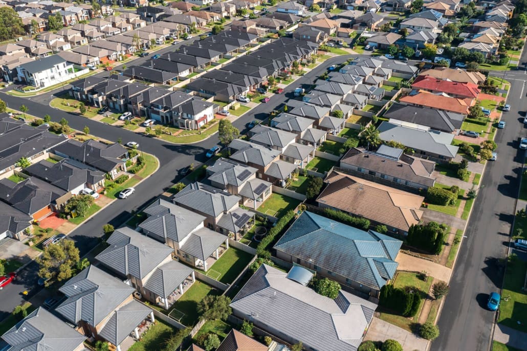 Aerial view of a suburban neighborhood with rows of modern houses, neatly arranged streets, green lawns, and trees lining the roads on a sunny day—ideal for insights from Property Valuers Sydney.