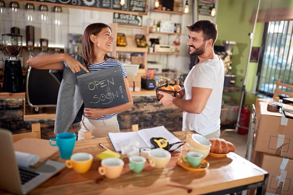 A smiling woman holds a chalkboard sign that says "OPEN SOON!" while a man with a basket of oranges looks at her in their cozy café, where mugs and pastries line the counter—perfectly capturing the spirit behind Small Business Valuations.