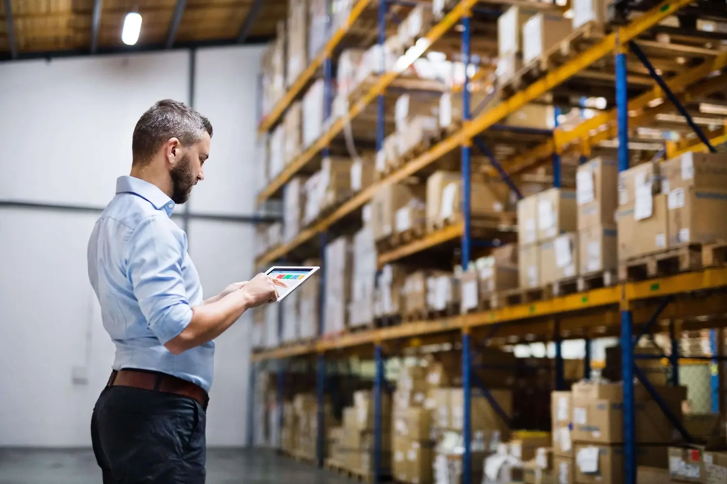 Stocktake Services Brisbane, A man in a blue shirt stands in a warehouse, holding a tablet and reviewing shelves filled with stacked cardboard boxes—a scene illustrating Stocktake Services Brisbane.
