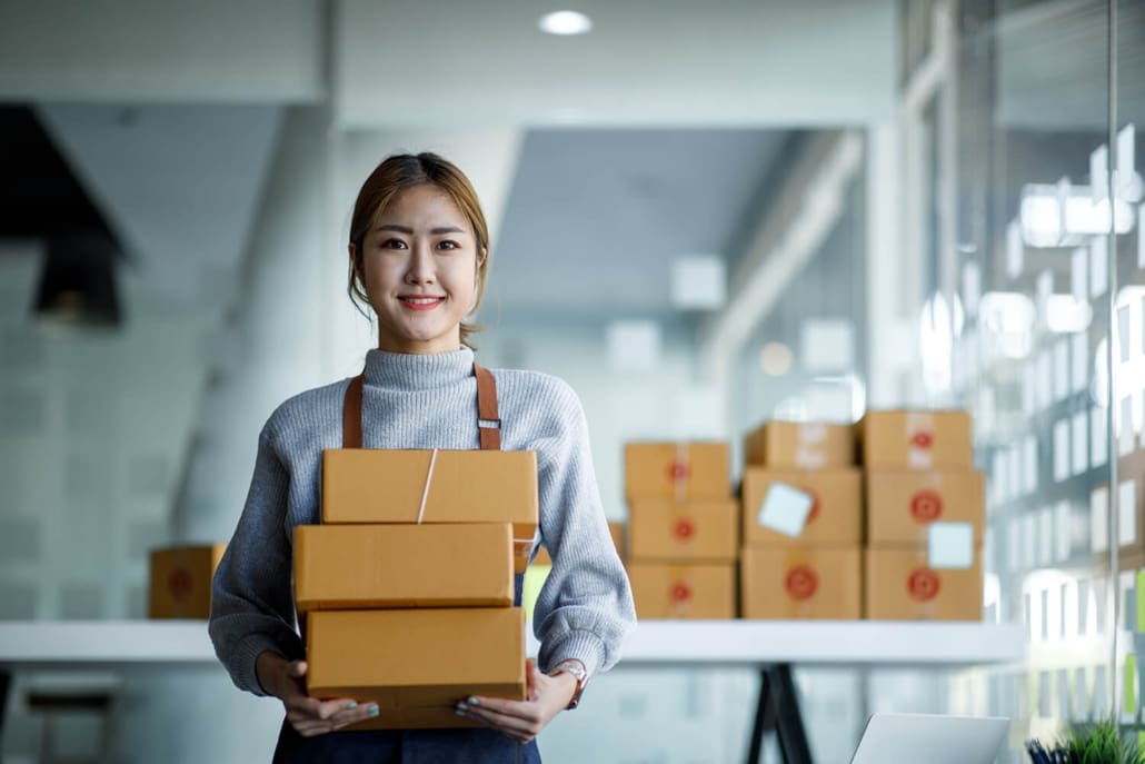 Stocktake Sydney - A woman wearing a gray sweater and apron smiles while holding three stacked cardboard boxes. In the background, more boxes rest on a white table in a bright, modern office space during a Stocktake Sydney event.